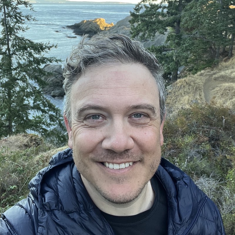Jason Klug, smiling in a puffy jacket on a rocky Salish Sea shoreline, evergreens and water behind him.